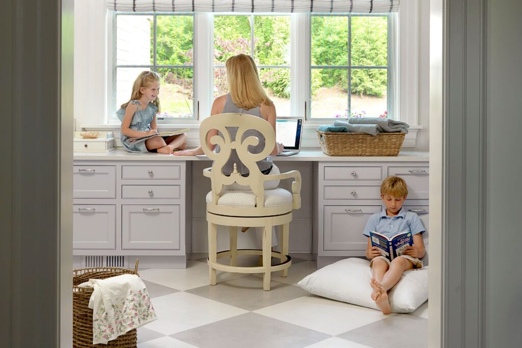 Woman sits at a laptop at a bright built‑in desk, with a girl on the window ledge drawing beside her and a boy reading on the floor nearby.