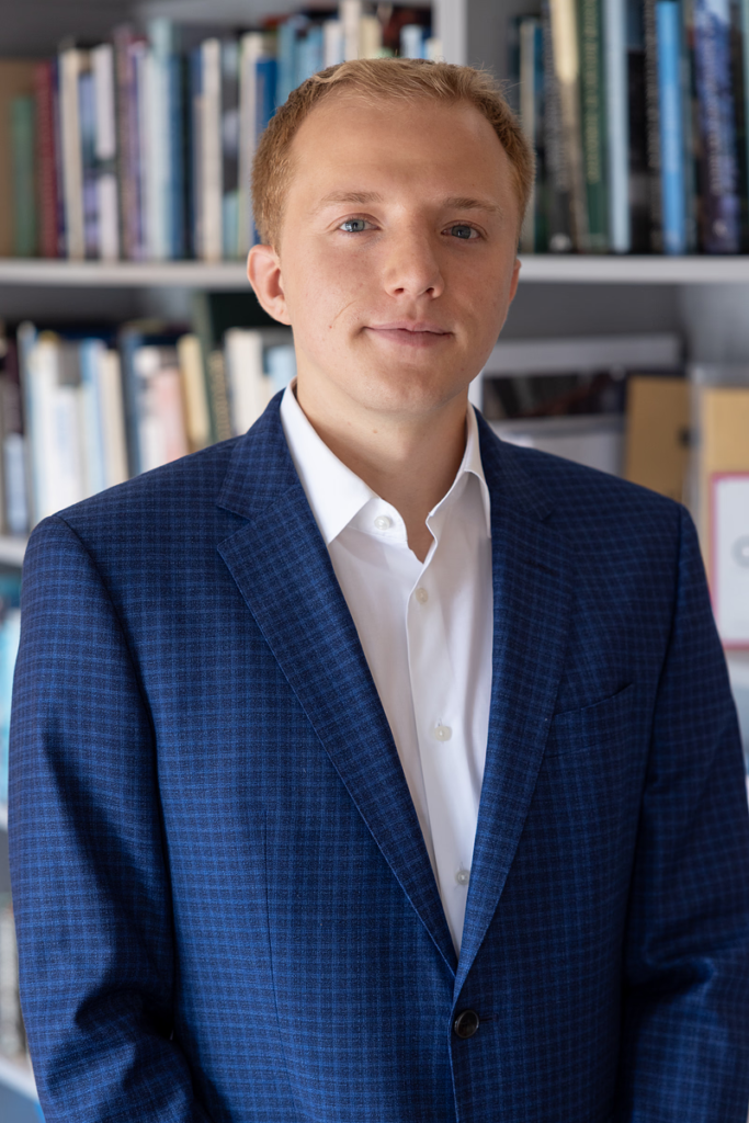 Portrait of a young man in a blue blazer and white shirt, standing in a library with shelves of books behind him, looking at the camera.